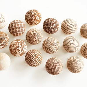 Peut inclure: Une collection de 15 boutons en bois avec différents motifs géométriques et floraux gravés à la surface. Les boutons sont tous de couleur brun clair et sont disposés en demi-cercle sur un fond blanc.
