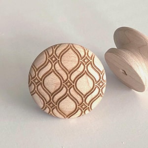 May include: Two round wooden knobs with a light tan natural wood grain finish. The front-facing knob features a deeply engraved, repeating geometric pattern of interlocking curved lines and small diamond shapes across its circular surface.