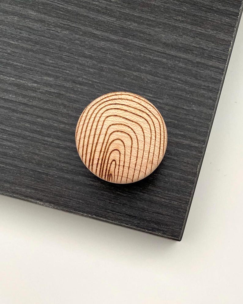 May include: A round, light-colored wooden cabinet knob with a concentric, curved grain pattern. The knob is set against a dark gray, textured surface, suggesting a modern or minimalist design aesthetic. The knob is likely for a drawer or cabinet.