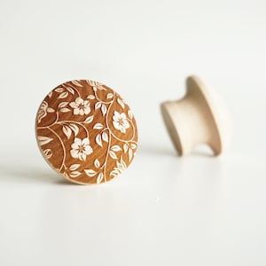 Peut inclure: Deux boutons de meuble en bois. L'un est rond avec un motif floral sculpté à la surface. L'autre est un bouton en bois simple et non fini. Les boutons sont brun clair et reposent sur une surface blanche.