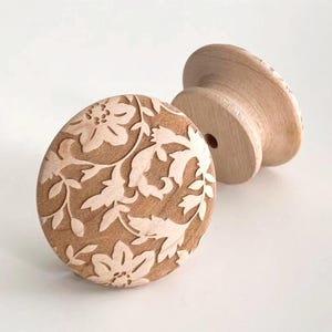 May include: Two unfinished wooden cabinet knobs. One knob features a detailed floral design carved into its round face. The other knob is a simple, unfinished wooden knob. The knobs are light brown in color and are set against a white background.