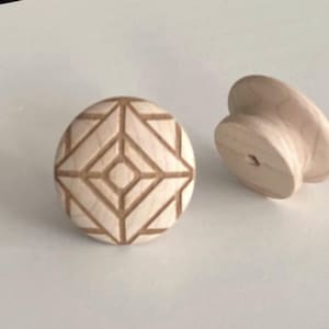 Puede incluir: Dos pomos de armario de madera sin terminar. Uno es redondo con un diseño geométrico tallado. El otro tiene forma de hongo. Los pomos son de madera de color claro y están sobre un fondo blanco.