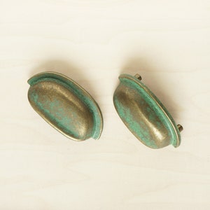 Könnte beinhalten: Zwei antike Messing-Schrankgriffe mit grüner Patina. Die Griffe sind oval geformt mit einer leichten Biegung und haben einen Used-Look.