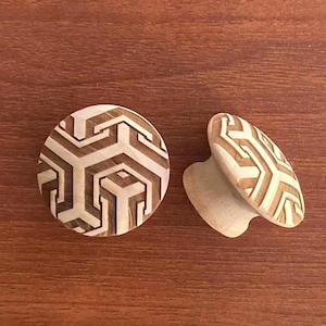 May include: Two round wooden cabinet knobs featuring a light and dark brown geometric pattern. The front-facing knob displays a repeating hexagonal design, while the side-view knob shows the cylindrical base and the continuation of the pattern.