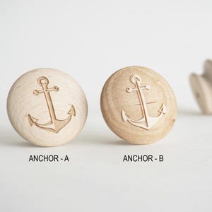 Puede incluir: Pomos de armario de madera con diseño de ancla. Se muestran dos pomos redondos, uno más claro y otro más oscuro, con un ancla tallada en la superficie. El texto "ANCHOR - A" y "ANCHOR - B" está debajo de los pomos. Un tercer pomo está en el fondo.