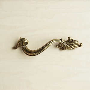 May include: A decorative brass drawer pull with a scroll design. The pull has a curved shape and intricate details.