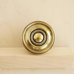 May include: A circular antique brass cabinet knob with a detailed concentric design. The knob has a central raised circle, surrounded by ridges and a decorative outer ring. The hardware is displayed on a light-coloured wooden surface.
