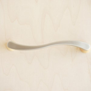 Op de afbeelding: Een zilveren kastgreep met een gebogen ontwerp en gouden eindkappen.