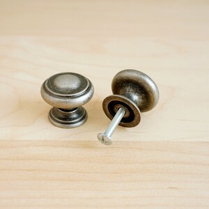 May include: Two silver cabinet knobs with a round, slightly domed top and a raised ring around the edge. One knob is attached to a screw.