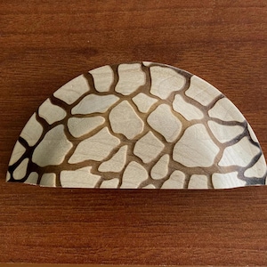 May include: A half-circle wooden cabinet pull with a light wood base and a pattern of irregular, light-colored shapes outlined in dark brown. The pull is set against a dark brown wooden surface.