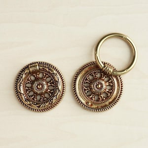 May include: Two ornate gold-toned metal drawer pulls. One pull is a round knob with a decorative floral design. The other pull is a ring with a decorative floral design.