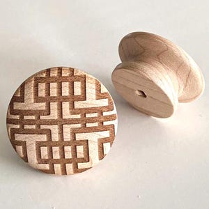 Puede incluir: Dos pomos de armario de madera sin terminar. Uno es redondo con un patrón geométrico grabado. El otro es un pomo curvo y macizo. Ambos están hechos de madera de color claro y están sobre un fondo blanco.