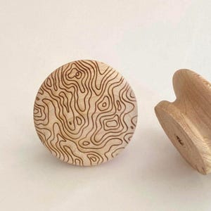 Könnte beinhalten: Zwei runde Holzknöpfe mit einer hellbraunen, natürlichen Holzmaserung. Der nach vorne gerichtete Knopf zeigt ein dunkelbraun graviertes topografisches Kartenmuster, das aus unregelmäßigen, fließenden Höhenlinien besteht.