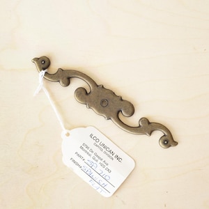 Könnte beinhalten: Ein verziertes Metall-Dekorstück in Goldton mit einem geschwungenen Design. Die Beschläge sind an einem weißen Etikett mit dem Text "ILCO UNICAN INC. CAPITOL DIVISION 5795 De Gaspé Ave Montreal, Que H25 2X3 PART # SP710 FINISH SPANISH FIT" befestigt.