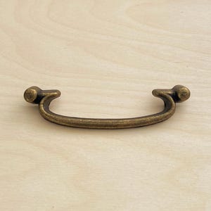 May include: A curved drawer pull handle with a distressed antique brass finish. The hardware features a central arched bar that connects to two rounded mounting points on either side, creating a smooth, continuous shape for furniture attachment.