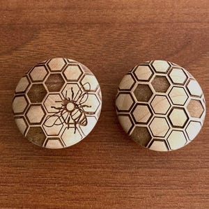 May include: Two round wooden decorative pieces with a honeycomb pattern. One piece features a detailed etched bee design within the honeycomb. The other piece has a honeycomb pattern. The pieces are light brown and sit on a brown wooden surface.