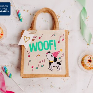 May include: A small brown hessian tote bag with a white handle. The bag features a cartoon dog wearing a party hat and the words "Woof!" in green lettering. The bag is surrounded by confetti and other party decorations.