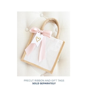 May include: A small, white burlap gift bag with a pink ribbon and a white gift tag with a gold heart. The bag is sitting on a light brown, textured surface.