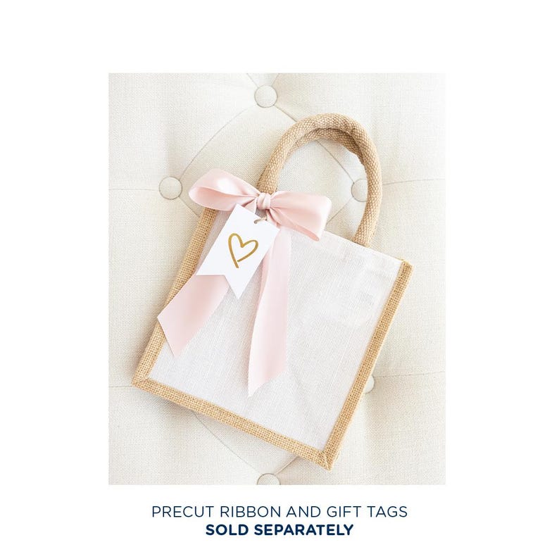 May include: A small, white burlap gift bag with a pink ribbon and a white gift tag with a gold heart. The bag is sitting on a light brown, textured surface.