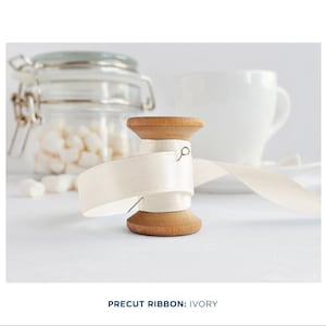 May include: A wooden spool of ivory satin ribbon with a pin holding the ribbon in place. The ribbon is partially unwound and the spool is sitting on a white surface.