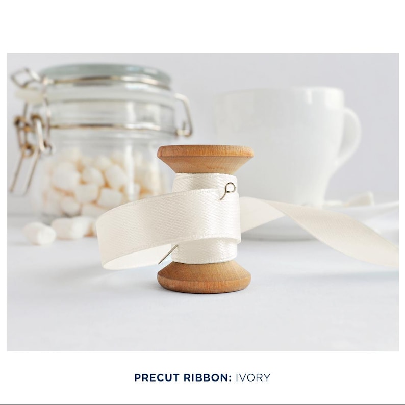 May include: A wooden spool of ivory satin ribbon with a pin holding the ribbon in place. The ribbon is partially unwound and the spool is sitting on a white surface.