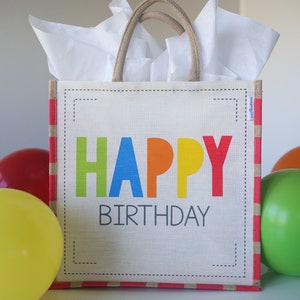 May include: A white gift bag with red and white stripes and a jute handle. The bag has a white label with the words "HAPPY BIRTHDAY" in colorful letters.