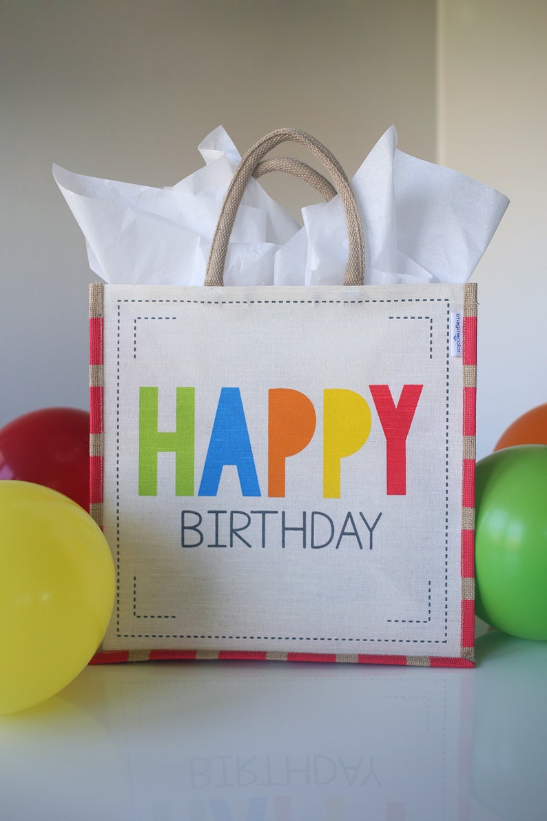May include: A white gift bag with red and white stripes and a jute handle. The bag has a white label with the words "HAPPY BIRTHDAY" in colorful letters.