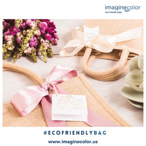 May include: Eco-friendly jute tote bags with pink satin ribbons and tags. One bag has a cream-colored bow. The bags have jute handles and are decorated with floral arrangements. The image includes the text "#ECOFRIENDLYBAG" and "www.imaginecolor.us".