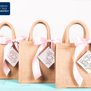 May include: Three small burlap gift bags with pink satin ribbon bows and white gift tags that say "Merci gracias thank you" in gold lettering.