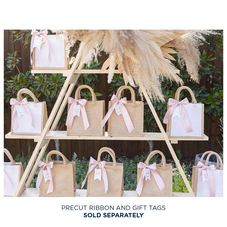 May include: A set of nine small burlap gift bags with pink ribbon and gold gift tags. The bags are displayed on a wooden shelf with a rustic background.