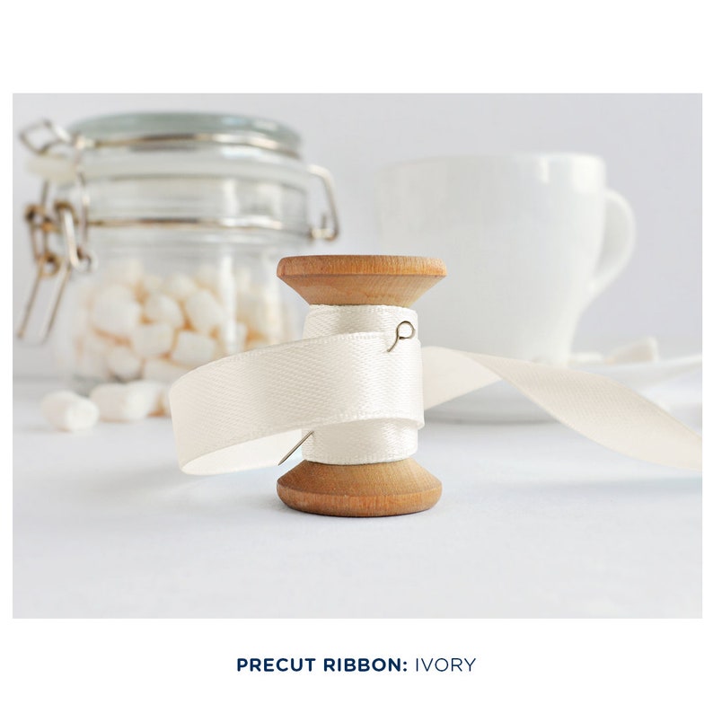May include: A spool of ivory satin ribbon wound around a wooden spool. The ribbon is partially unwound and a pin is sticking out of the spool.