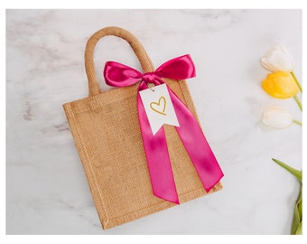 Può includere: Una piccola borsa di juta marrone con un nastro di raso rosa legato a fiocco. Un'etichetta regalo bianca con un cuore dorato è attaccata al nastro. La borsa è su una superficie di marmo bianco.