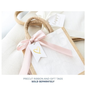 May include: A white burlap tote bag with a pink ribbon and a white gift tag with a gold heart. The bag is tied with a pink ribbon and has a gold heart on the tag. The bag is sitting on a white surface.