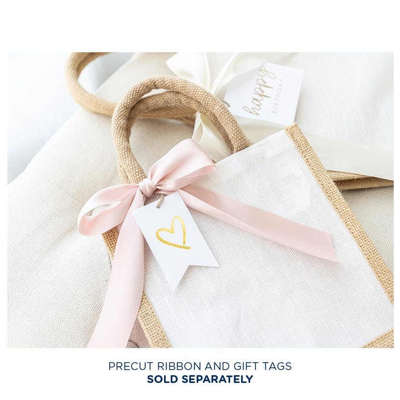 May include: A white burlap tote bag with a pink ribbon and a white gift tag with a gold heart. The bag is tied with a pink ribbon and has a gold heart on the tag. The bag is sitting on a white surface.