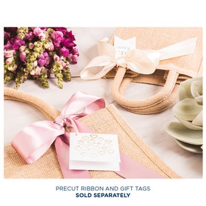 May include: Two burlap gift bags with pink and cream ribbon bows. One bag has a white gift tag with gold hearts. The text "Precut ribbon and gift tags sold separately" is at the bottom of the image.