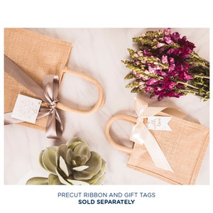 May include: Two small burlap gift bags with handles, one with a silver ribbon and a tag that says "with love", the other with a beige ribbon and a tag with a heart design. The bags are on a white surface with a succulent plant and a bouquet of purple flowers.