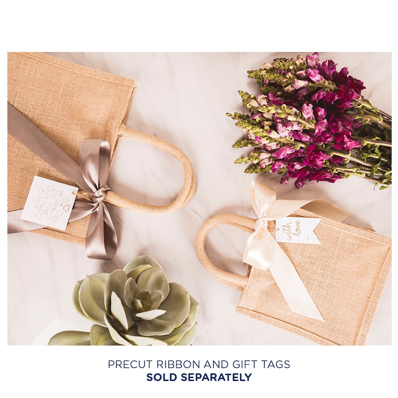 May include: Two small burlap gift bags with handles, one with a silver ribbon and a tag that says "with love", the other with a beige ribbon and a tag with a heart design. The bags are on a white surface with a succulent plant and a bouquet of purple flowers.