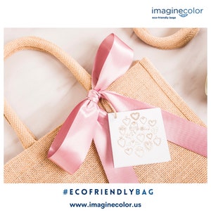 May include: A natural burlap tote bag with pink ribbon tied in a bow. The bag has a tag attached with a gold heart design and the text "#ECOFRIENDLYBAG www.imaginecolor.us".