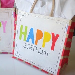 May include: A white reusable tote bag with red and beige stripes. The bag has a  "Happy Birthday" message with colorful letters.