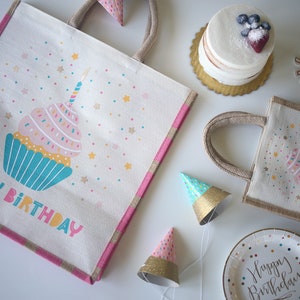 May include: Two white burlap tote bags with pink and blue cupcake designs and the text "Happy Birthday". The bags are surrounded by party supplies, including a cake, party hats, and a paper plate.