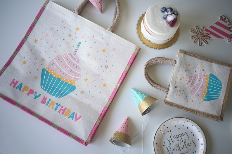 May include: Two white burlap tote bags with pink and blue cupcake designs and the text "Happy Birthday". The bags are surrounded by party supplies, including a cake, party hats, and a paper plate.