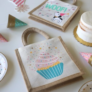 May include: A set of party favours for a birthday party. The favours include a small burlap bag with a cupcake design, a small burlap bag with a dog design, and a few party hats with glitter and confetti.