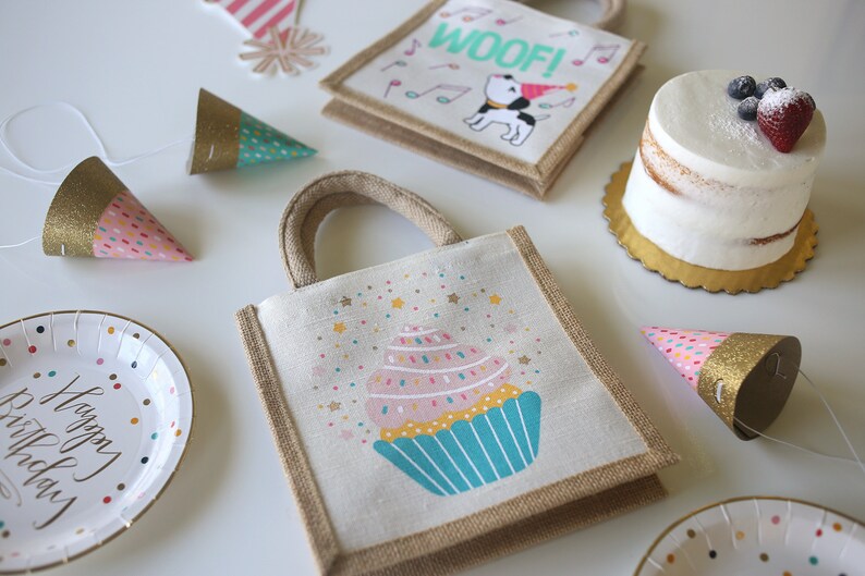 May include: A set of party favours for a birthday party. The favours include a small burlap bag with a cupcake design, a small burlap bag with a dog design, and a few party hats with glitter and confetti.