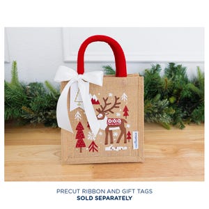May include: A small burlap gift bag with a red handle and a white bow. The bag features a Christmas design with a reindeer, trees, and mountains in red and white. The bag is sitting on a wooden surface.