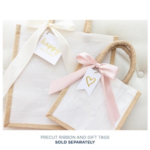 May include: Two white burlap gift bags with brown trim. The larger bag has a white ribbon and a gift tag that says "happy birthday". The smaller bag has a pink ribbon and a gift tag with a gold heart.