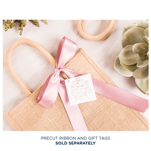 May include: A natural burlap tote bag with woven handles, tied with a light pink satin ribbon. A white tag with gold heart designs hangs from the ribbon. The text "PRECUT RIBBON AND GIFT TAGS SOLD SEPARATELY" is at the bottom.