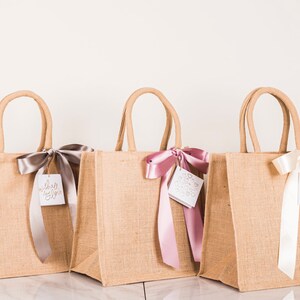 May include: Three burlap tote bags with matching handles. Each bag is adorned with a satin ribbon and a tag. The ribbons are in silver, pink, and white. The tags have the words "with all my love" and other designs.