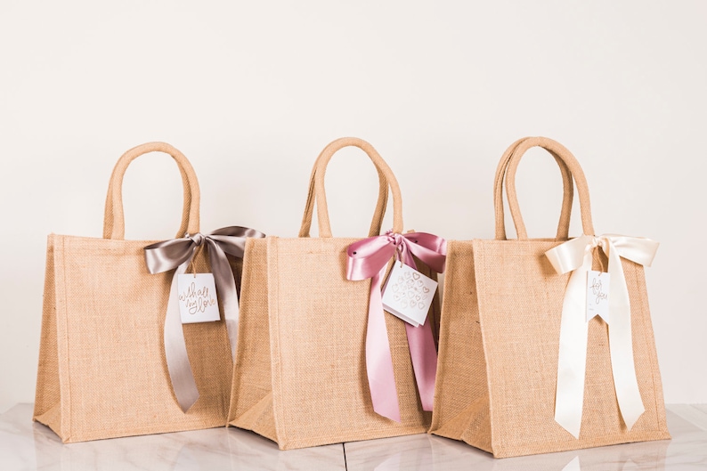 May include: Three burlap tote bags with matching handles. Each bag is adorned with a satin ribbon and a tag. The ribbons are in silver, pink, and white. The tags have the words "with all my love" and other designs.