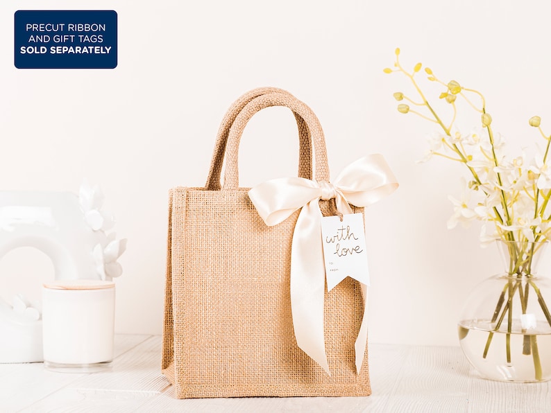 May include: A brown burlap tote bag with a white satin ribbon tied around the handles. The bag has a white gift tag attached to the ribbon that says "with love".