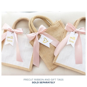 May include: Three burlap tote bags with white fabric fronts and woven handles. Each bag is adorned with a pink ribbon and a white tag with gold text or a gold heart. The tags read "with love".